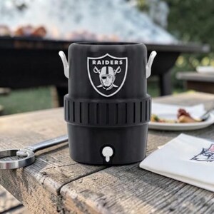 May include: Black insulated beverage cooler with the Raiders logo, featuring a white emblem with a football player's face. The cooler has a spigot and two silver handles. A bottle opener and a napkin are on a wooden table.