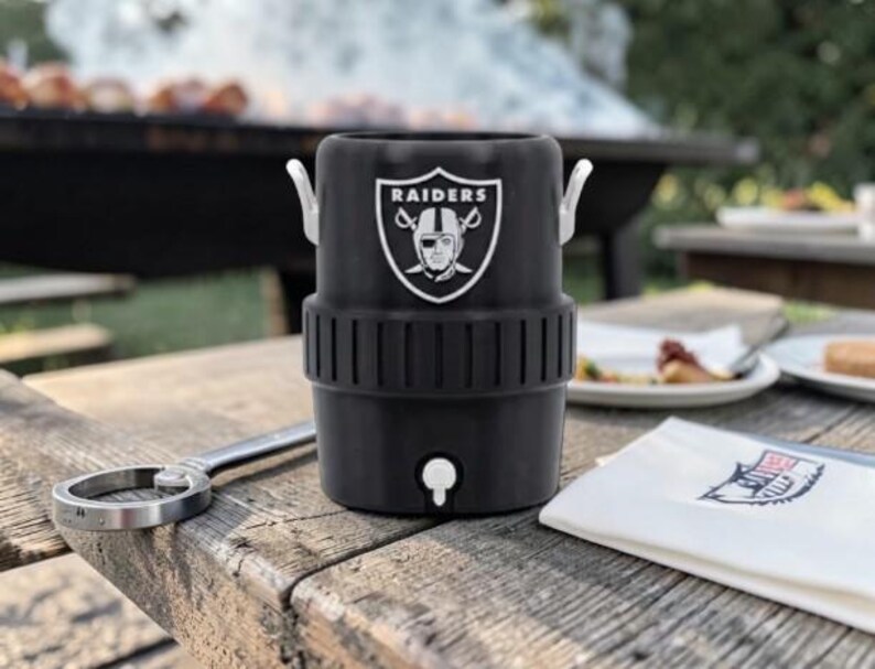 May include: Black insulated beverage cooler with the Raiders logo, featuring a white emblem with a football player's face. The cooler has a spigot and two silver handles. A bottle opener and a napkin are on a wooden table.