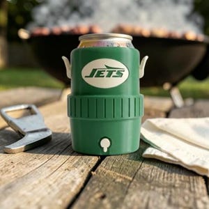 May include: A green insulated can cooler with the New York Jets logo, a silver bottle opener, and a white cloth on a wooden surface. A barbecue grill is in the background.