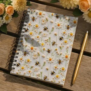 May include: A spiral-bound notebook with a white cover featuring embroidered daisies and bees. A gold pen rests beside the notebook on a wooden surface, with floral arrangements in the background. The notebook's design includes yellow, white, and black elements.