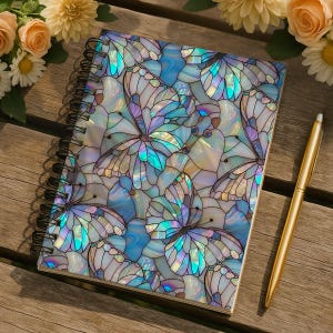 May include: Spiral-bound notebook with a cover featuring a stained-glass butterfly design in shades of blue, white, and iridescent colors. A gold pen rests beside the notebook on a wooden surface. Floral accents are in the background.