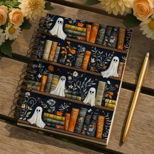 May include: Spiral-bound notebook with a dark blue cover featuring a bookshelf design with books and floral accents. White ghost figures are placed on the shelves. A gold pen rests beside the notebook on a wooden surface.