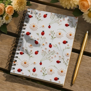 May include: A spiral-bound notebook with a white cover featuring a pattern of red ladybugs, white daisies, and green foliage. A gold pen rests on a wooden surface next to the notebook. Floral arrangements of yellow and peach flowers are in the background.