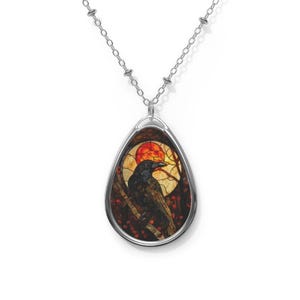 May include: Silver-toned pendant necklace featuring a teardrop-shaped pendant with a stained-glass design. The design depicts a black raven perched on a branch against a backdrop of a red sun and autumn foliage. The chain is a delicate silver color.