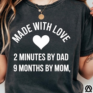 May include: Dark gray t-shirt with the text "MADE WITH LOVE" arched above a white heart. Below the heart, the text reads "2 MINUTES BY DAD 9 MONTHS BY MOM,". The shirt has a vintage, worn look.