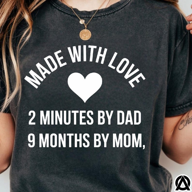 May include: Dark gray t-shirt with the text "MADE WITH LOVE" arched above a white heart. Below the heart, the text reads "2 MINUTES BY DAD 9 MONTHS BY MOM,". The shirt has a vintage, worn look.