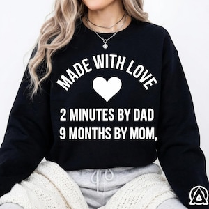May include: Black sweatshirt with the text "MADE WITH LOVE" above a white heart, and the text "2 MINUTES BY DAD 9 MONTHS BY MOM,". The sweatshirt is worn by a person with blonde hair.