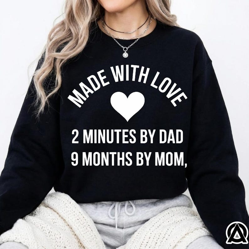 May include: Black sweatshirt with the text "MADE WITH LOVE" above a white heart, and the text "2 MINUTES BY DAD 9 MONTHS BY MOM,". The sweatshirt is worn by a person with blonde hair.