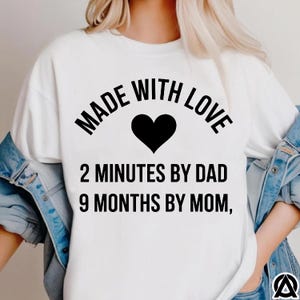 May include: White t-shirt with the text "MADE WITH LOVE" above a black heart. Below the heart, the text reads "2 MINUTES BY DAD 9 MONTHS BY MOM,". The shirt is worn with a denim jacket.