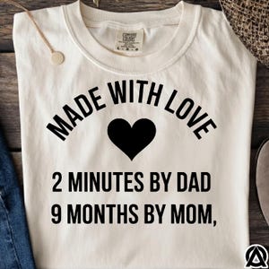 May include: Cream-colored t-shirt with the text "MADE WITH LOVE" above a black heart. Below the heart, the shirt reads "2 MINUTES BY DAD 9 MONTHS BY MOM,". The shirt has a crew neck and short sleeves.