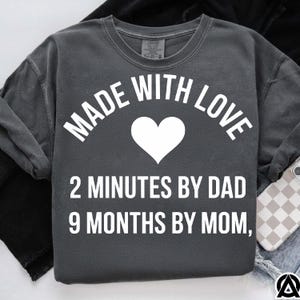 May include: Dark gray sweatshirt with the text "MADE WITH LOVE" above a white heart. Below the heart, the text reads "2 MINUTES BY DAD 9 MONTHS BY MOM". The sweatshirt is a comfortable, casual style.