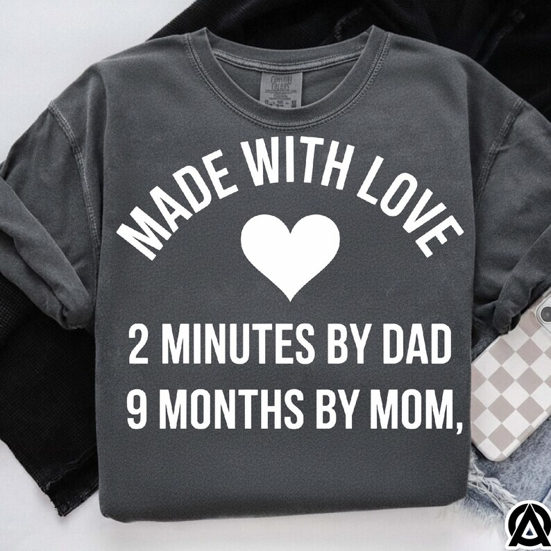 May include: Dark gray sweatshirt with the text "MADE WITH LOVE" above a white heart. Below the heart, the text reads "2 MINUTES BY DAD 9 MONTHS BY MOM". The sweatshirt is a comfortable, casual style.
