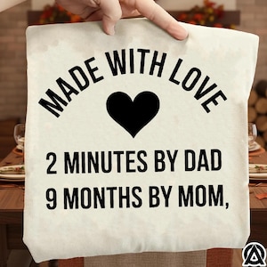 May include: A cream-colored pillow with the text "MADE WITH LOVE" in black, a black heart, and the phrases "2 MINUTES BY DAD" and "9 MONTHS BY MOM," also in black. The pillow is being held up by a hand.