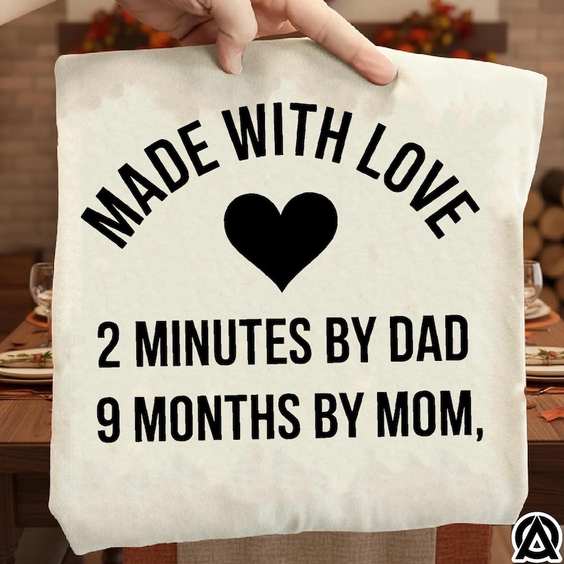 May include: A cream-colored pillow with the text "MADE WITH LOVE" in black, a black heart, and the phrases "2 MINUTES BY DAD" and "9 MONTHS BY MOM," also in black. The pillow is being held up by a hand.