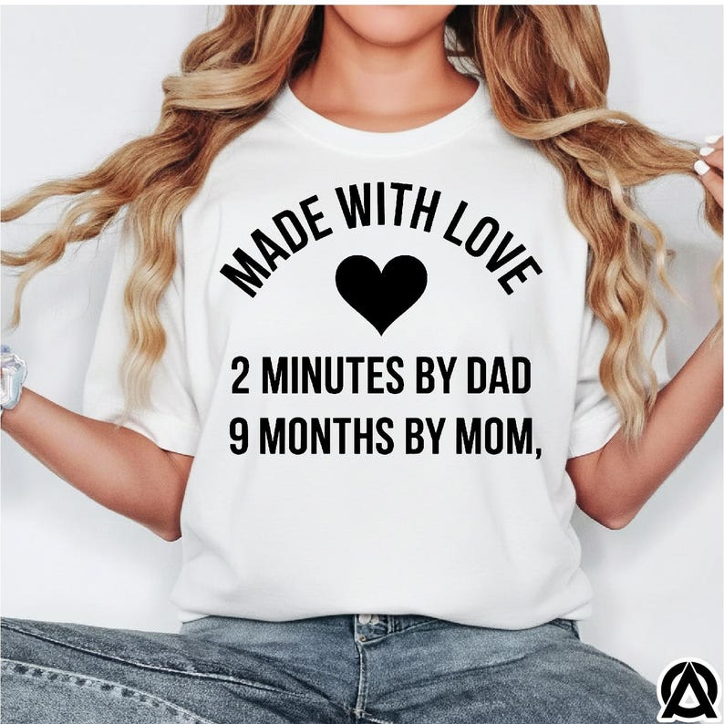 May include: White t-shirt with the text "MADE WITH LOVE" above a black heart. Below the heart, the text reads "2 MINUTES BY DAD 9 MONTHS BY MOM,". The shirt is a casual style.