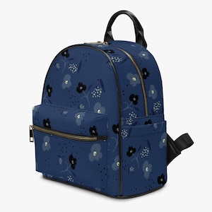 Puede incluir: Una mochila azul marino con un estampado floral de flores negras y azul claro y motas blancas. La mochila tiene un asa negra, correas negras y cremalleras doradas. La bolsa tiene un bolsillo frontal y dos bolsillos laterales.