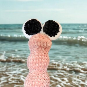 May include: A whimsical, handmade plush creature with a pink body and large, round eyes. The eyes are black with white borders. The plush toy is set against a blurred background of a beach and ocean.
