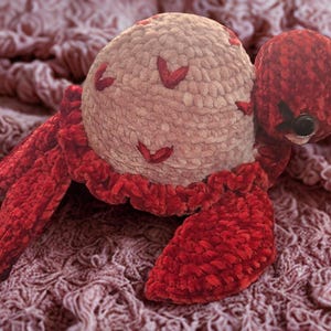 May include: A handmade plush crab toy with a light beige body and red accents. The crab has red legs, claws, and a ruffled trim around the body. The crab's body has red heart-shaped details.