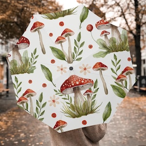 May include: White umbrella with a repeating pattern of red-capped mushrooms with white spots, green leaves, and small flowers. The umbrella is open, displaying the design. The background is blurred.