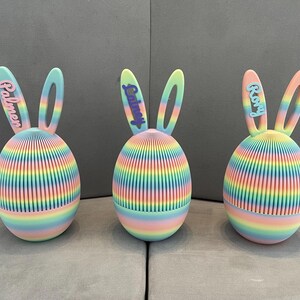 May include: Three decorative Easter egg ornaments in a rainbow colour scheme, each with bunny ears. The eggs have vertical stripes and the names Palmer, Quincy, and Roxy on the ears. They are displayed on a grey surface.