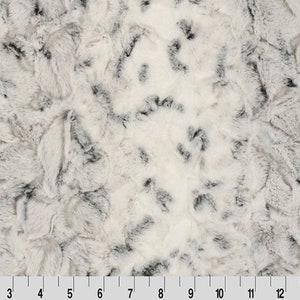 May include: Close-up of a faux fur fabric with a cream-colored base and irregular black markings. The fabric has a soft, textured appearance, with a ruler along the bottom edge showing inch measurements.