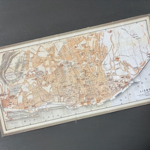 May include: A vintage map of Lisbon, Portugal, with a detailed street layout in shades of brown and beige. The map is framed with a light-colored border and includes the word "LISBOA" in the lower right corner.
