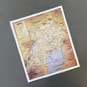 May include: A rectangular map of Uganda, with surrounding countries labeled, including Sudan, Kenya, and Tanzania. The map is framed with a white border and features a detailed depiction of the region's geography, including Lake Victoria.