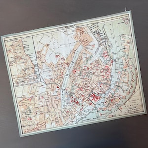 May include: A vintage map of Copenhagen, Denmark, with a detailed layout of streets, waterways, and landmarks. The map features a cream-colored background with red and brown details, framed by a light green border. The word "KOPENHAGEN" is printed at the top.