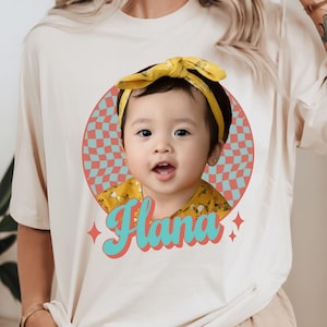 May include: Cream-colored t-shirt featuring a graphic of a child with a yellow headband and shirt. The child is framed by a retro circle with a checkered pattern. The name "Hana" is written in teal script below the child.