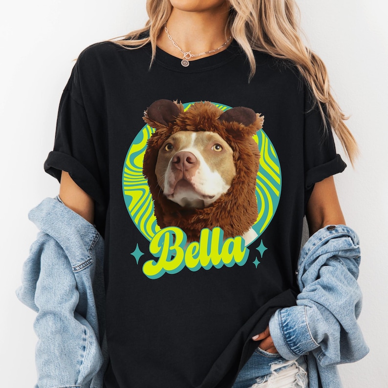May include: Black t-shirt featuring a dog wearing a brown lion's mane costume. The dog's face is centered within a green and blue circular design. The name "Bella" is written in yellow script below the dog.