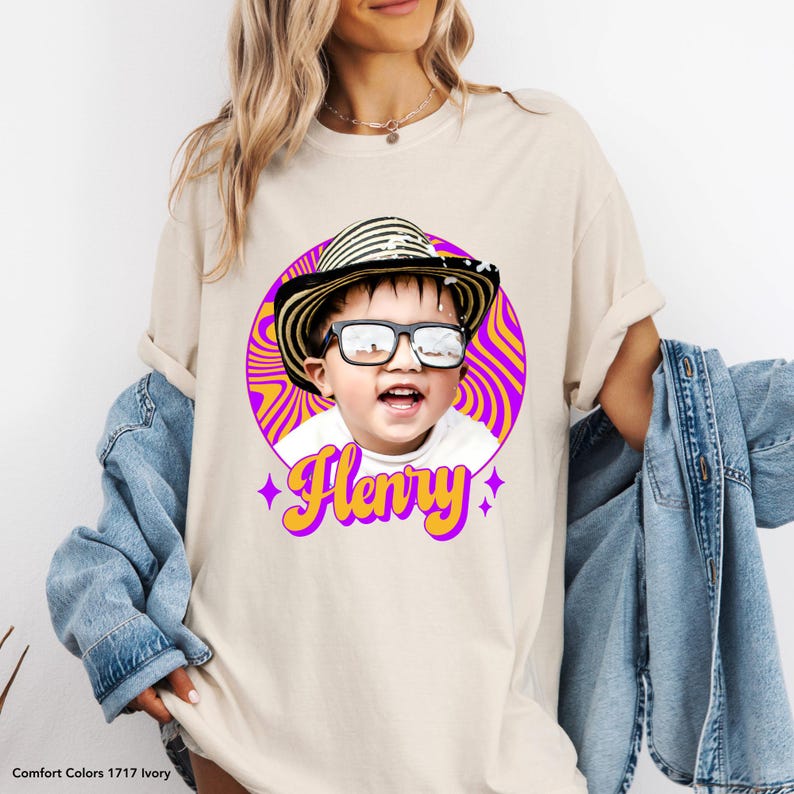May include: Ivory-colored t-shirt featuring a graphic of a child wearing sunglasses and a striped hat, set against a purple and orange swirl background. The name "Henry" is written in yellow with purple star accents.