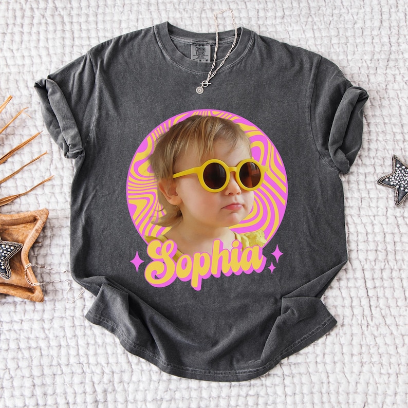 May include: Dark gray t-shirt featuring a graphic of a child wearing yellow sunglasses, set against a pink and yellow swirl background. The name "Sophia" is written in pink and yellow script below the image.