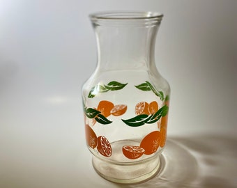 May include: A clear glass pitcher with a citrus design. The pitcher has a wide base and a narrow neck. The design features green leaves and orange slices.