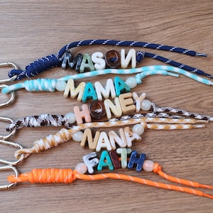 May include: Five keychains featuring silver-toned metal clasps and colorful paracord lanyards. Each lanyard holds letter beads spelling MASON, MAMA, HONEY, NANA, and FAITH, accented with round beads in various colors, textures, and patterns.