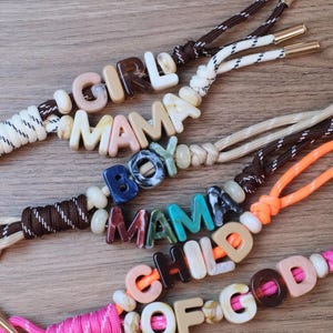 May include: Four braided cord bracelets featuring acrylic letter beads. The bracelets display the words GIRL MAMA, BOY MAMA, CHILD, and OF GOD in various colors including brown, white, blue, green, orange, and pink with metallic end caps.