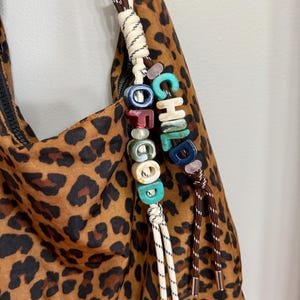 May include: A leopard print bag featuring a decorative charm made of two knotted cords. The charm includes multi-colored, marbled, and solid-colored beads spelling out the word CHILDHOOD, accented with small light pink spherical beads.