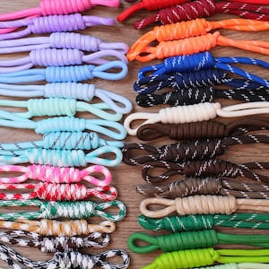 May include: A collection of multi-colored paracord zipper pulls arranged in two columns. The cords feature various solid colors and patterns, including neon, pastel, and dark shades, each tied with a barrel knot to form a loop at one end.