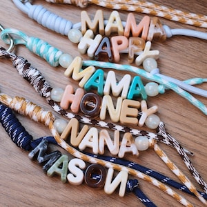 May include: A collection of four wristlet keychains made from braided paracord in various colors including light blue, teal, brown, and navy. Each keychain features custom acrylic letter beads spelling MAMA HAPPY, MAMA HONEY, NANA, and MASON.