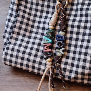 May include: A bag charm featuring two knotted cords attached to a gold-toned metal clip. One cord is beige with beads spelling MAMA, and the other is dark brown with beads spelling BOY. Both cords end in gold-toned metal caps and hang from a bag.