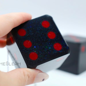A close up view of two holographic black car die with red dot pips with a linked silver chain. The die is being held by a model which shows the die fits in her palm.