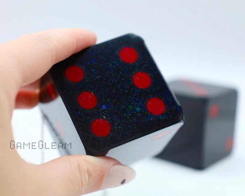 A close up view of two holographic black car die with red dot pips with a linked silver chain. The die is being held by a model which shows the die fits in her palm.