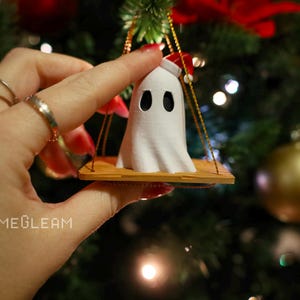 A close up view of a white Santa ghost on a brown swing ornament hanging on a holiday Christmas tree with lights in the background. A model has her hand holding the ornament.