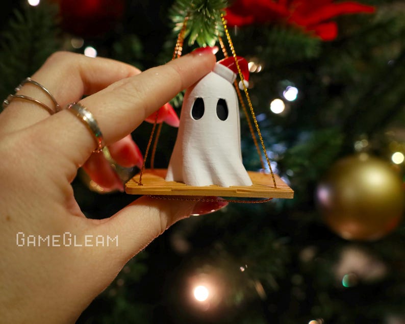 A close up view of a white Santa ghost on a brown swing ornament hanging on a holiday Christmas tree with lights in the background. A model has her hand holding the ornament.