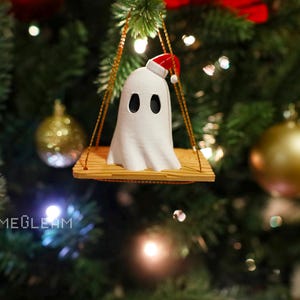 A close up view of a white Santa ghost on a brown swing ornament hanging on a holiday Christmas tree with lights in the background.