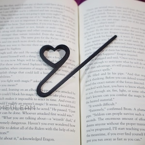 Pode incluir: Um marcador de página de metal preto em forma de coração com uma ponta afiada. O marcador de página está aberto em um livro com páginas brancas e texto preto. O texto na página diz "Como se luta com magia?" e "O que você quer dizer?"
