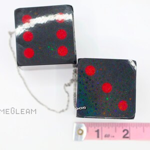 A close up top-down view of two holographic black car dice with red dot pips and a linked silver chain laying on a white surface. Next to them is a pink measuring tape showing each die is two inches long.