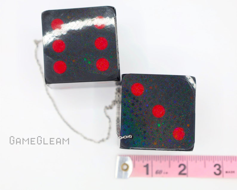 A close up top-down view of two holographic black car dice with red dot pips and a linked silver chain laying on a white surface. Next to them is a pink measuring tape showing each die is two inches long.