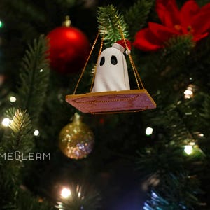 A bottoms up view of a white Santa ghost on a brown swing ornament hanging on a holiday Christmas tree with lights in the background.