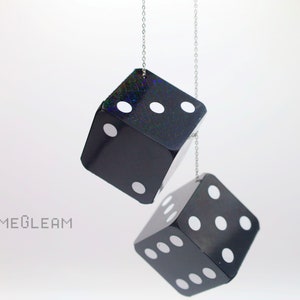 A close up side view of two holographic black car dice with white dot pips and a linked silver chain hanging over a white surface.
