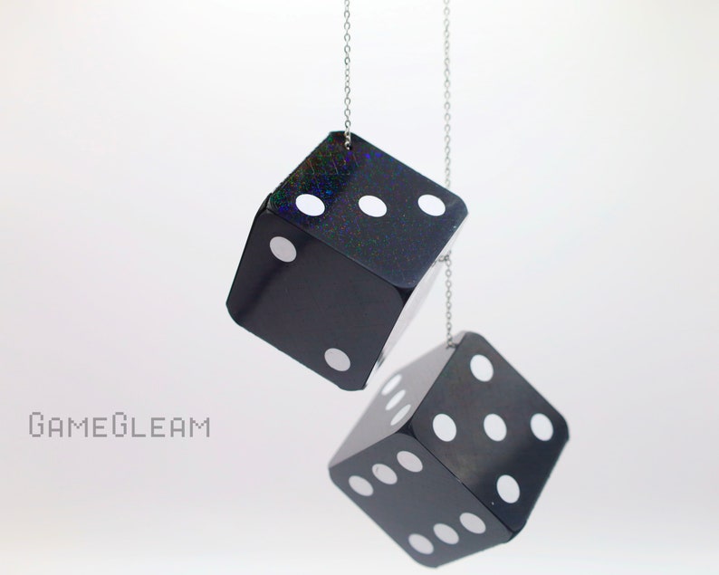 A close up side view of two holographic black car dice with white dot pips and a linked silver chain hanging over a white surface.
